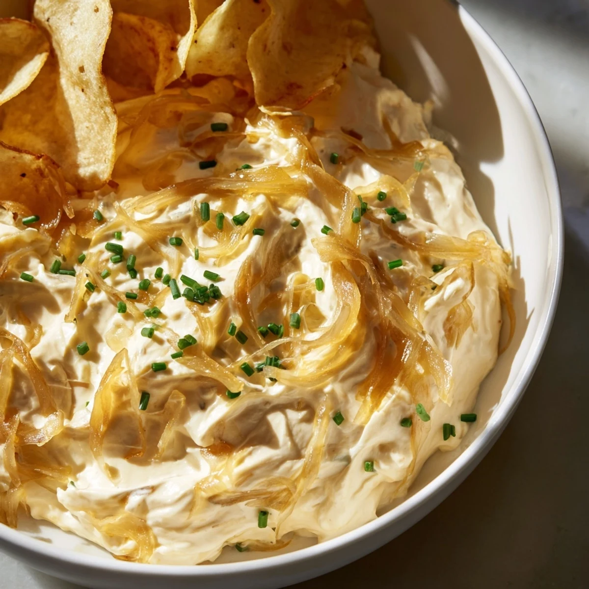 Close-up of golden, caramelized onion dip topped with extra chives, served alongside crunchy thick-cut potato chips.